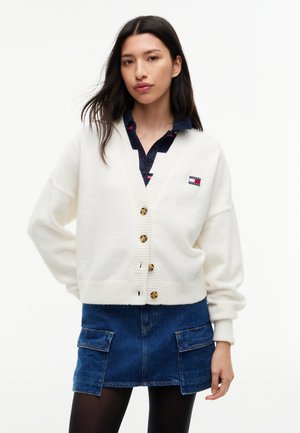 Mujer con cárdigan blanco abotonado, camisa azul marino, falda vaquera con bolsillos y medias negras, de pie mirando hacia adelante con una mano en la cadera.