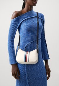 Vrouw in een blauw geribd jurk met één schouder, met een witte crossbodytas met verticale strepen in blauw, rood en zwart en een zwarte band.