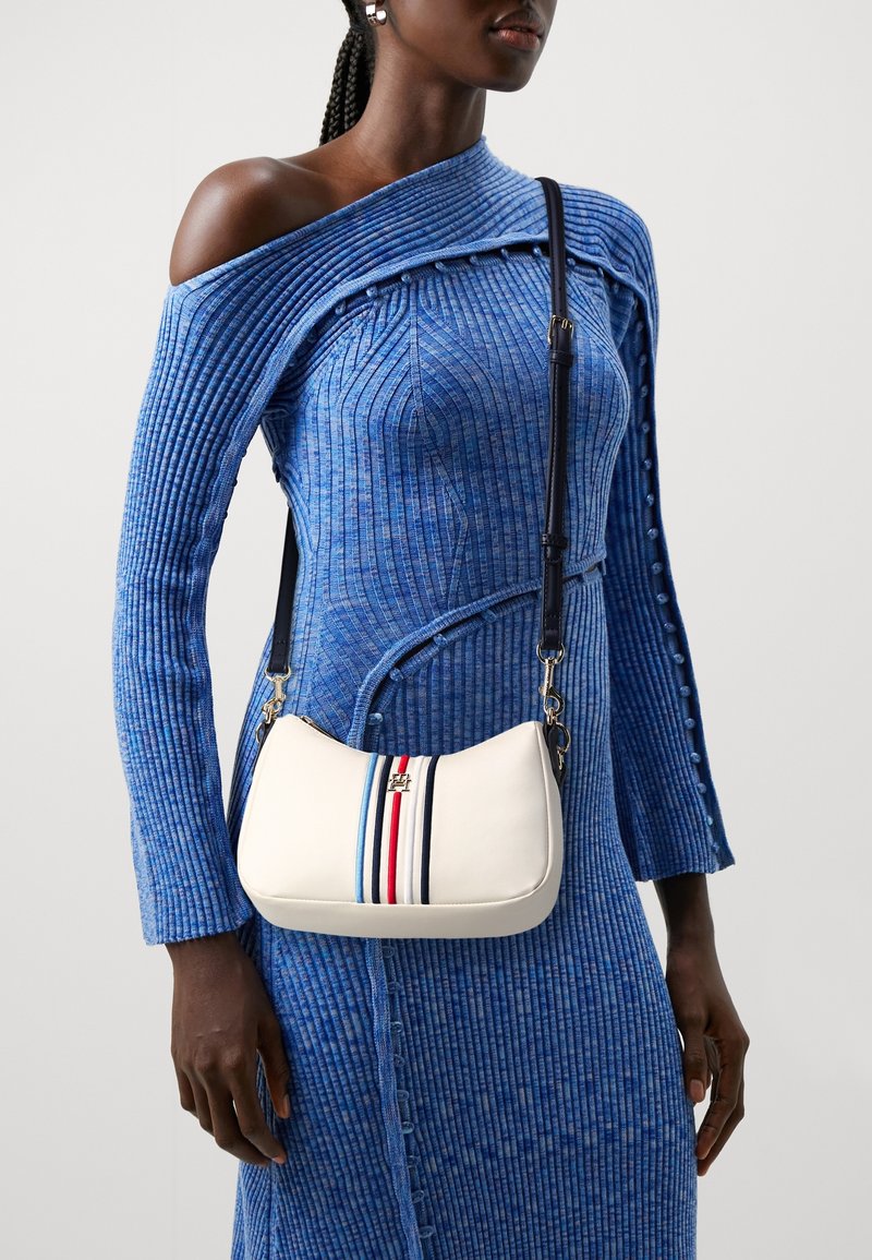 Vrouw in een blauw geribd jurk met één schouder, met een witte crossbodytas met verticale strepen in blauw, rood en zwart en een zwarte band.