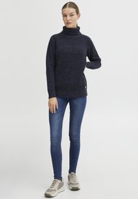 Navy knitted sweater with a high roll neck, textured fabric, ribbed cuffs, and hem, paired with fitted blue jeans and light sneakers.