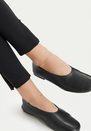 Person wearing black fitted pants and black leather slip-on shoes with delicate gold anklets on both ankles.