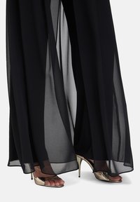 Un tissu noir, transparent, tombant jusqu'au sol drape les jambes, associé à des sandales à talons hauts en métal doré avec un design à bout ouvert.