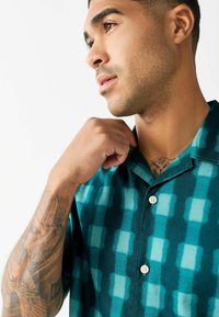 Cămașă cu mânecă scurtă în nuanțe de teal și albastru închis, având un model în grilă, cu deschidere pe butoane și un guler relaxat. Textură a țesăturii netedă.