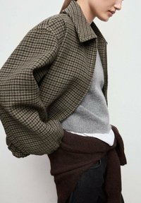 Chaqueta corta de cuadro houndstooth marrón con un cuello estructurado, combinada con un suéter gris y un envoltura de punto marrón. Presenta mangas voluminosas.
