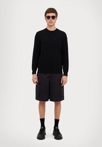 Maglione nero a maglia con texture geometrica, abbinato a pantaloni cargo scuri fino al ginocchio, calzini neri e sneakers nere. Occhiali da sole marroni.