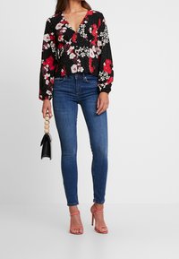 Blusa nera floreale con scollatura a V profonda, maniche lunghe e motivi di fiori rossi e bianchi. Abbinata a jeans skinny blu e sandali con cinturini rosa.