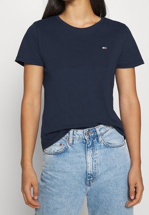 Vrouw draagt een marineblauw T-shirt met korte mouwen en een klein logo op de borst, gecombineerd met hoge lichtblauwe jeans, tegen een witte achtergrond.