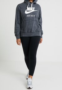 Grå Nike hoodie med vit logotyp, har en framficka och dragsnören. Kombinerad med svarta leggings och vita sportskor.