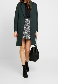 Zelený dlouhý kabát s klopovým límcem, dvěma předními kapsami, nošený přes černý top a vzorovanou sukni. Černá kabelka a kotníkové boty doplňují vzhled.