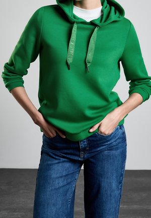 Persoon draagt een felgroene hoodie met trekkoorden en opgerolde mouwen, gecombineerd met een blauwe spijkerbroek, handen in de zakken.