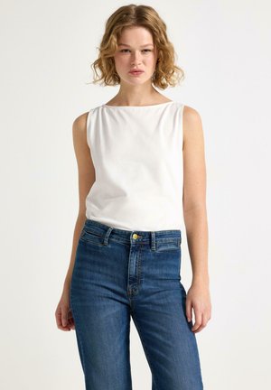 Kvinde med krøllet blondt hår iført en hvid ærmeløs top og blå højtaljede jeans, stående mod en hvid baggrund.