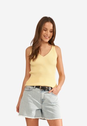 Femme souriante, portant un haut jaune côtelé sans manches et un short en jean clair avec une ceinture noire, posant avec une main dans la poche.