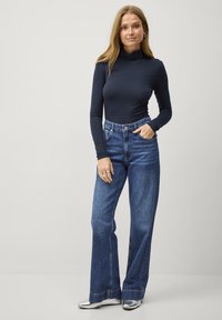 Mørkeblå langærmet turtleneck-top parret med højtaljede, blå flared jeans og metallisk sølvfarvede sko; model står med en hånd i lommen.