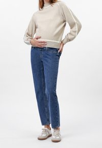 Osoba v béžovém svetru a modrých džínech, stojící s jednou rukou na břiše a druhou v kapse, na bílém pozadí.