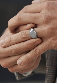 Sølvring med en glatt, oval topp som hviler på en hånd, med polert metallfinish. Hender er delvis sammenfoldet og viser naturlig hudtekstur.