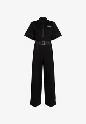 Črn kombinezon s kratkimi rokavi, z zadrgo in kontrastnim belim šivom. Ima širok pas in ohlapne hlače.