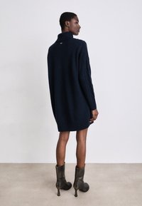 Ναυτικό πλεκτό φόρεμα με ψηλό γιακά, ραμμένο στρίφωμα και λεπτομερείς μακριές μανίκες. Συνδυάζεται με μαύρες γόβες με ψηλό τακούνι.