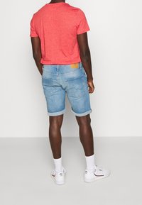 Červené tričko s krátkým rukávem vyrobené z měkké tkaniny je spárováno se světle modrými denimovými šortkami. Šortky mají ohrnuté manžety a na zadní kapse béžovou nášivku.