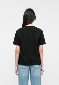 T-shirt de algodão preto com mangas curtas, corte descontraído e bainha reta. Visto de trás, mostrando o detalhe da etiqueta azul na gola.
