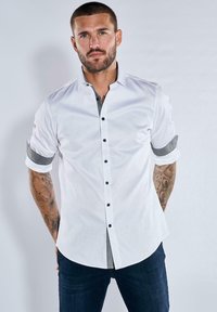 Weißes Langarmhemd mit schwarzen Knöpfen, grau-karierten Akzenten an den hochgekrempelten Ärmeln, kombiniert mit dunklen Denim-Jeans. Glatte Stofftextur.
