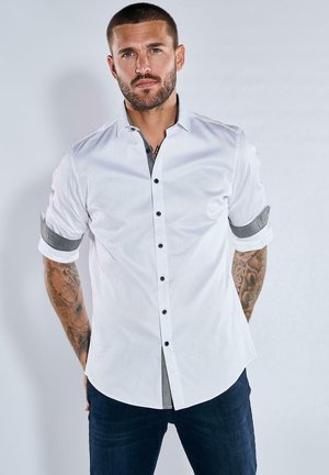 Weißes Langarmhemd mit schwarzen Knöpfen, grau-karierten Akzenten an den hochgekrempelten Ärmeln, kombiniert mit dunklen Denim-Jeans. Glatte Stofftextur.