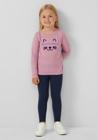 Maglietta a maniche lunghe rosa con grafica di un volto di gatto, abbinata a leggings blu scuro e sneakers bianche. Design semplice, vestibilità casual per bambini.