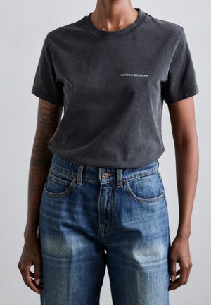 Osoba w wyblakłej czarnej koszulce Victoria Beckham włożonej do wysokich niebieskich dżinsów, z widocznymi tatuażami na ramionach.