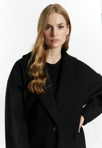 Černý oversize kabát vyrobený z vlněné směsi, s šálovým límcem, jedním knoflíkovým zapínáním a mírně texturovanou látkou.