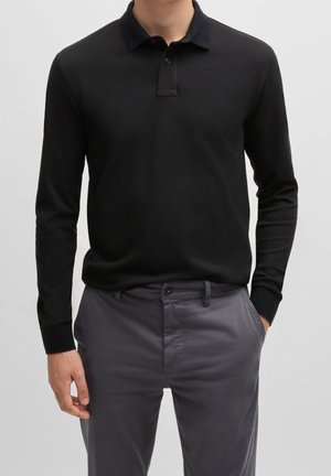 Schwarz langärmliges Polohemd mit einem kontrastierenden dunklen Kragen, gefertigt aus glattem Stoff. Kombiniert mit grauen, leichten Hosen mit Fronttaschen.