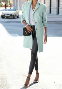 Frau, die auf der Straße geht und einen mintgrünen Mantel, einen weißen Pullover, schwarze Lederhosen, eine schwarze Handtasche und schwarze Open-Toe-Highheels trägt.