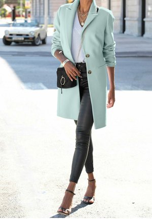 Frau, die auf der Straße geht und einen mintgrünen Mantel, einen weißen Pullover, schwarze Lederhosen, eine schwarze Handtasche und schwarze Open-Toe-Highheels trägt.
