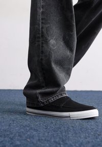 Sorte sneakers med teksturert finish og hvite gummisåler, kombinert med mørkegrå jeans som har et subtilt mønster.