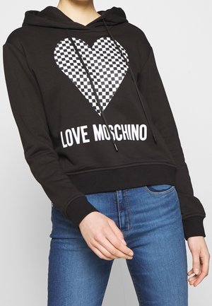 Osoba nosząca czarną bluzę z kapturem z wzorem w kratkę w kształcie serca oraz tekstem "Love Moschino", połączoną z niebieskimi dżinsami na jednolitym tle.