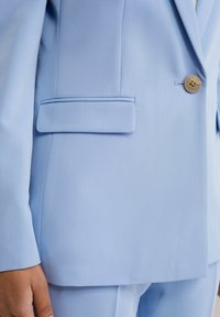 Ljusblå kavaj i ett mjukt tyg. Har en snedskärning på reversen, en enkel knäppning och en sidficka med en slät finish.