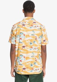 Kortärmad, gul skjorta med knappar som har ett tropiskt mönster med palmer, röda blommor och segelbåtar, tillverkad av lättviktsmaterial.