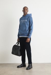 Felpa di cotone blu con logo "HILFIGER", borsa nera, stivali Chelsea neri e pantaloni scuri. Texture liscia, vestibilità casual e design minimalista.