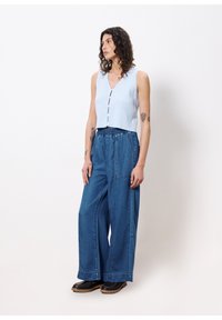 Top sans manches côtelé bleu clair avec des boutons noirs et un pantalon en denim bleu à jambes larges avec une taille élastique et des poches. Chaussures noires.