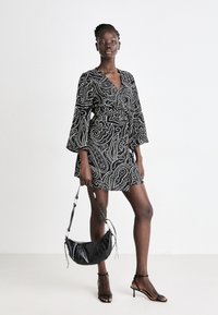 Robe en imprimé paisley noir et blanc avec des manches en cloche, design enveloppant et ourlet évasé. Le mannequin tient un sac à main en cuir noir et porte des talons à lanières.