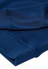 Tumši zils džemperis ar gludu tekstūru, ar ribotām piedurknēm un iepresētu logotipa detaļu. Salocīts ar tīrām malām.