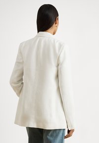 Λευκό λινό σακάκι με ραμμένη σιλουέτα, πέτο με εγκοπές και μανσέτες με τρία κουμπιά, σε συνδυασμό με ανοιχτό μπλε τζιν παντελόνι.