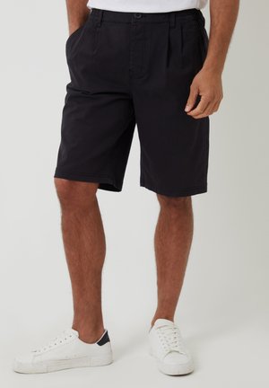 Pantalones cortos negros de algodón, con un corte holgado, bolsillos frontales y cierre de botón. Combinados con zapatillas blancas.