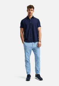 Camisa de manga corta azul marino con tela texturizada, cierre de botones, combinada con pantalones ceñidos de color azul claro. El calzado son zapatillas oscuras.