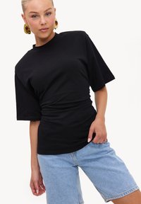 Femme portant de grandes boucles d'oreilles en or, un t-shirt noir à manches courtes et un short en denim bleu clair, posant avec une main dans la poche.