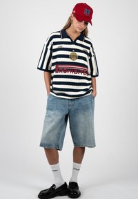 Persona que lleva una gorra roja, una camiseta polo de rayas amplia con el texto "PROHIBITED", pantalones cortos de mezclilla hasta la rodilla, calcetines blancos y mocasines negros.