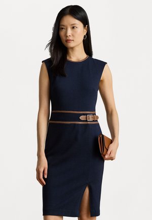 Marineblauwe mouwloze jurk met een getailleerd ontwerp, voorzien van een bruine riem in de taille en een split aan de voorkant. Houdt een kleine bruine clutch vast.