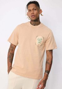 T-shirt beige à manches courtes en coton, avec une poche ornée d'un graphisme botanique vert et d'un contour rond et texturé.