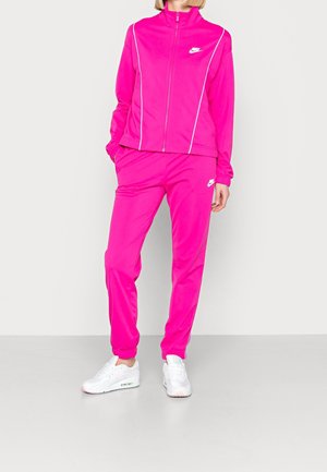 Personne portant un survêtement Nike rose vif avec des liserés blancs et des baskets Nike blanches, debout devant un fond clair uni.