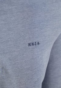 Gestructureerde lichtblauwe stof met een subtiel rasterpatroon. Bevat een donkerblauw logo "NZLA" geborduurd op het oppervlak.