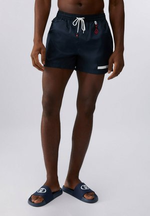 Uomo con pantaloncini da bagno blu navy con coulisse bianca e sandali navy, in piedi davanti a uno sfondo grigio semplice.