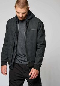 Veste bomber noire pour homme avec une surface texturée, un devant zippé et une capuche amovible, portée sur une chemise gris foncé.
